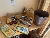 Six shot glasses with decorative designs, including a swallow, bowling pin, and others arranged on a wooden table with coasters, tongs, and metal planter