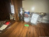 Photo showing various glass and ceramic beer mugs on a wooden shelf, including a large glass mug with embossed circles and a ceramic stein with winter horse scene.