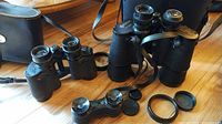 Photo showing Tasco 8x30 binoculars, Carl Wetzlar 10x50 binoculars, and vintage opera glasses with black textured finishes, focus wheels, and protective caps on a wooden floor.