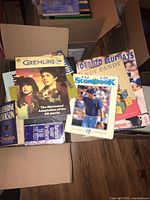 Photo showing an open cardboard box with various books inside, including Gremlins film adaptation, Toronto Blue Jays sports book, a sports scorebook, and other novels.