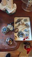 Brooches and pins on wooden surface with a ceramic base object visible at top left, showing overall lot contents including some larger and smaller brooches with stones and floral decorations.