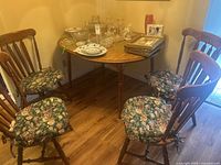 Four wooden spindle-back chairs arranged around a small round wooden table, each with a floral seat cushion, placed on hardwood floor.