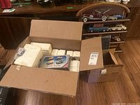 Open cardboard box with styrofoam packing containing boxed Classic Trucks set and other boxed model cars, wooden display shelf visible in background.