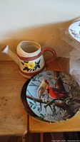 Ceramic watering can with a yellow flower design and a crack, next to three collector plates with birds on branches, placed on wooden table.