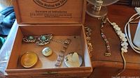 Wooden box open showing various jewellery pieces including bracelets, a compact mirror, and a pill box inside, along with bracelets/necklaces on the side