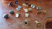 Overview of 10 sets of cufflinks on wooden surface including various shapes and colors, some with enamel and stones.