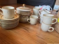 Set of 8 stoneware cups, 8 matching saucers, and a covered bowl displayed on a wooden table. Items have a cream base with brown accent bands and rustic finish.