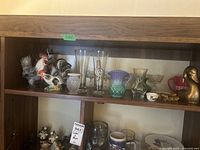 Shelf with assorted china rooster figurines, glass vases, glasses, mug, and brass duck figurine.