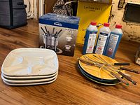 Photo showing the fondue set boxed, four white divided plates stacked, four yellow glazed round plates stacked, four wooden-handled forks, and three bottles of liquid fondue fuel.
