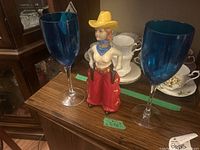Photo showing two tall blue plastic wine glasses and a painted cowboy figurine bottle on wooden furniture.