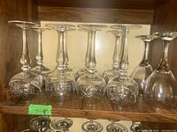 Eight crystal glasses with etched floral pattern, arranged upright showing decorative design along bowl and stem on a wooden shelf.