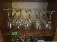Fifteen clear glass wine glasses on a wooden shelf, viewed from front showing the glasses upside down with visible stems and bowl shape.