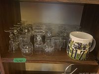 Shelf containing approximately twenty liquor and shot glasses, with a large decorative ceramic mug on the right side.