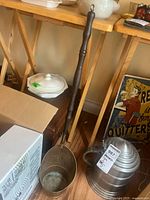 Full view of the metal ladle showing the wooden handle and deep bowl, leaning against a wall with surrounding furniture and decor.