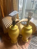 Photo showing two yellow ceramic table lamps, 17 inch each, with no shades, plugged in cords, standing side by side near window light.