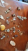 Mismatched jewelry assortment spread on wood surface including cufflinks, earrings, brooches, pendants, and pins with various metals and designs.