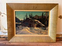 Framed winter scene painting showing log cabin with snow and pine trees, whole painting and frame visible