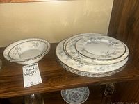 Four stacked plates and one soup bowl with floral Brides Choice pattern and silver trim shown on a wooden shelf