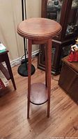 Full view of the tall oak plant stand showing the round top, three legs, and bottom support shelf on hardwood floor beside furniture.