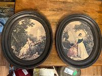 Pair of framed oval prints viewed from above on wooden surface showing entire frames and prints