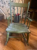 Front view of green painted child size wooden rocking chair showing the seat, armrests, spindle backrest, and rockers with visible scratches and paint chips.