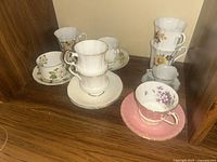 Full set of six teacups with saucers arranged on a wooden shelf, showing the variety of patterns and shapes.