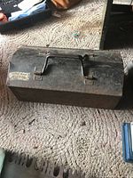 Front view of closed rectangular metal toolbox with visible handle and latch, showing worn black finish and rust spots, placed on carpet.