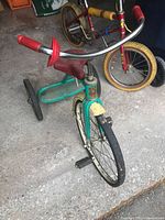 Front and side angle of vintage green Leader tricycle showing front wheel, handlebar with red grips, seat and footrest platform.