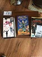 Photo of 3 VHS tapes on wooden surface: My Fair Lady, Peter Pan, Scrooge. Each has MaxSold sticker labels.