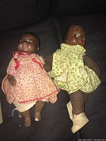 Two vintage dolls sitting on dark fabric, one in red floral dress and one in yellow floral dress with shoes and tights