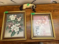 Two framed floral 3D prints and a ceramic cat figurine on a wooden surface. The left print shows white and pink blossoms, the right print shows pink lilies, both in gold-tone frames. The cream and brown ceramic cat figurine is resting with eyes closed.