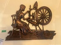 Close-up photo of metal wall art showing detailed figure of woman at spinning wheel with textured bronze surface.