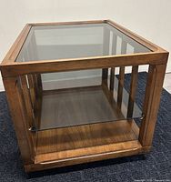 Square walnut end table with smoked glass top and vertical wood columns on two sides on a dark carpeted floor against white wall.