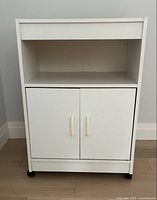 Front view of white microwave stand showing open top shelf, two-door cabinet below and caster wheels.