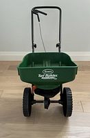 Front view of Scott's Turf Builder Mini Spreader showing green hopper with logo, black handle, and wheels.