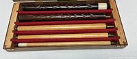 Top-down view of the 5 piece pool cue laid in carved wooden case with red felt lining. Shows carved dark wood sections and lighter wood sections with white ferrules and threaded joints.