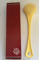 Yellow Avon bath brush with soft white bristles alongside the original red Avon box featuring a white floral design on a textured red background.