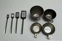 Top view of four stainless steel measuring spoons and four stainless steel Mary Ann measuring cups on a white surface.