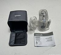 Photo showing the Waterpik water flosser device, power cord, instruction manual, and open zippered storage case on a white surface.