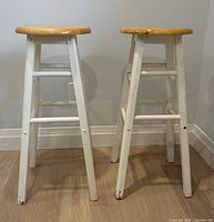 Two wooden bar stools with natural wood seats and white painted legs shown from front view on hardwood floor against light gray walls.