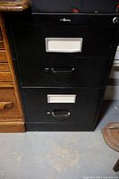 Front view of black 2 drawer metal filing cabinet with label holders and pull handles visible.