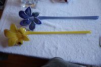 Two long-stem Murano glass flowers, one yellow and one blue, laying on a white cloth background.
