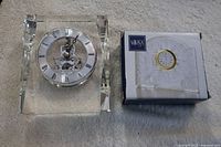 Two small desk clocks side by side on carpet. One clock is a transparent skeleton clock inside a clear crystal block, the other is boxed Mikasa crystal clock.