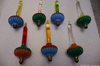 Eight vintage bubble lights arranged on white fabric, showing multicolored fluted bases with colored liquid inside glass tubes, various colors include blue, green, red bases and yellow, red, clear liquids.