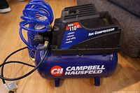 Campbell Hausfeld blue and black portable air compressor with coiled air hose and power cord on wooden floor