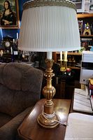 Full view of the large table lamp showing the gold-tone ornate base, cord, and white pleated silk lampshade with gold lace trim, on a wooden table beside soft seating and decorative items.