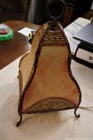 Side view of the leather henna Moroccan lamp showing the curved or tear-drop shape and hand-stitched seams with decorative patterns along edges. Lamp stands on three small curled metal feet with metal ring handle on top.