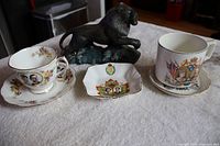 Five royal souvenir items shown on a cloth surface including two commemorative cups with saucers, a small commemorative dish, a hand-painted lion figurine, and a coffee mug