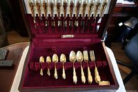 Cutlery set arranged inside the velvet-lined wooden box showing knives, forks, spoons and serving utensils with gold plated floral design handles.