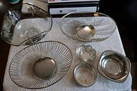 Overview of all bowls and dishes in lot on white cloth, showing glass bowl, silver plated wire bread bowls, small dishes, creamer, sugar bowl, and shaker.